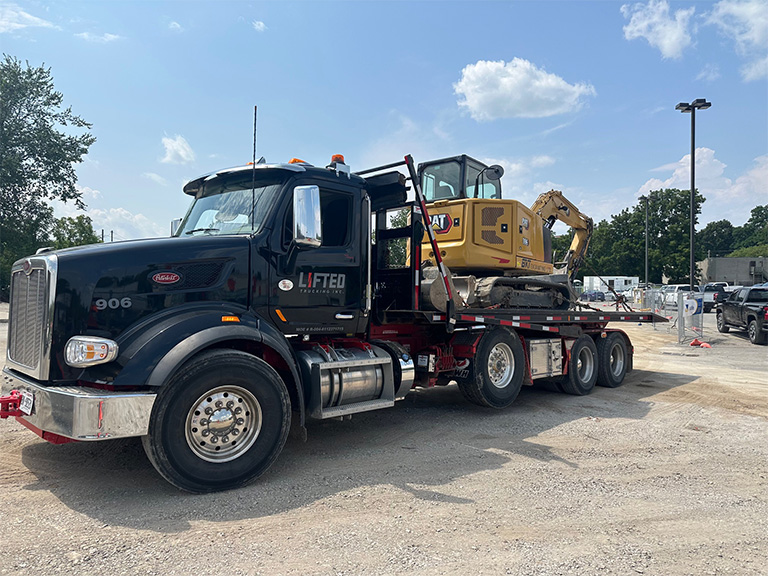 A photo of a triaxle flatbed truck with an excavator on the back.